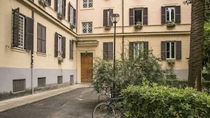 Une ruelle étroite où un vélo est garé près d’une haie, avec une porte en bois et un immeuble doté de nombreuses fenêtres et volets.