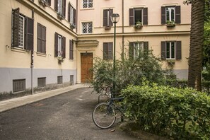 A narrow alleyway with a bicycle parked by a hedge, a wooden door, and a building with multiple windows and shutters.