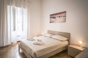 A bedroom with a wooden bed, white linens, a nightstand with a lamp, and a painting on the wall.