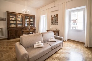 Una sala de estar con sofá, un aparador de madera, una araña, una mesa de comedor y una ventana.