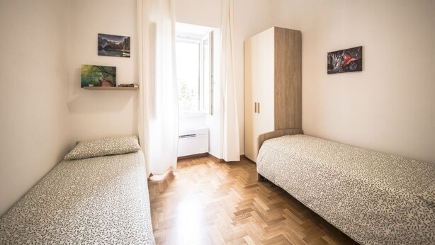 Une petite pièce bien éclairée, avec deux lits simples, une armoire en bois, une fenêtre avec des rideaux et des œuvres d’art accrochées au mur.