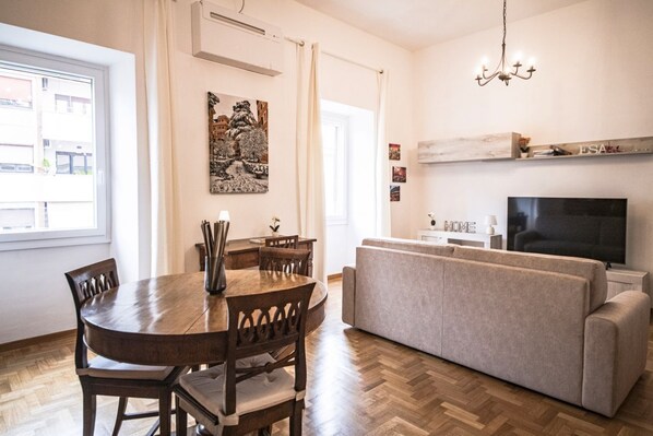 A modern living room with a sofa, a TV, a dining table with chairs, and a painting on the wall.