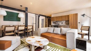 Un soggiorno moderno con divano, poltrone, tavolino da caffè e angolo cottura con pensili in legno e zoccolo in marmo.
