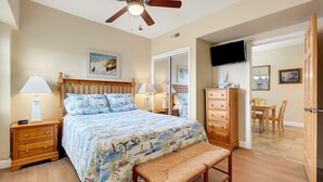 A bedroom with a bed, bedside tables, a wooden dresser, a bench, a ceiling fan, a TV, and a dining area with chairs and a table. - FFR201 My Happy Place by First Flight Rentals (Kill Devil Hills)