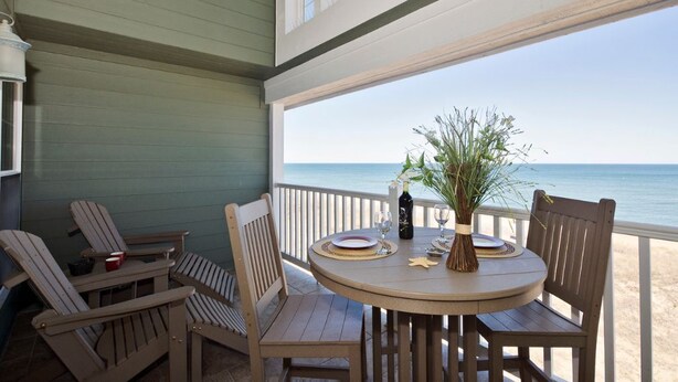 A round wooden table set for two with chairs, a bottle, and a vase of greenery on a balcony overlooking the ocean. - FFR314 - Just Wright by First Flight Rentals (Kill Devil Hills)