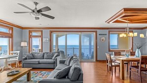 A spacious living area with a large gray sofa, a dining table with chairs, and a view of the ocean through large windows. - WV24 Kiteboarding Paradise by First Flight Rentals (Rodanthe)