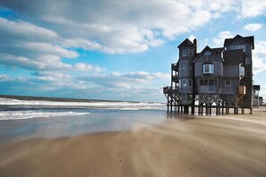Um edifício costeiro com vários andares e varandas, localizado numa praia de areia com o oceano ao fundo.