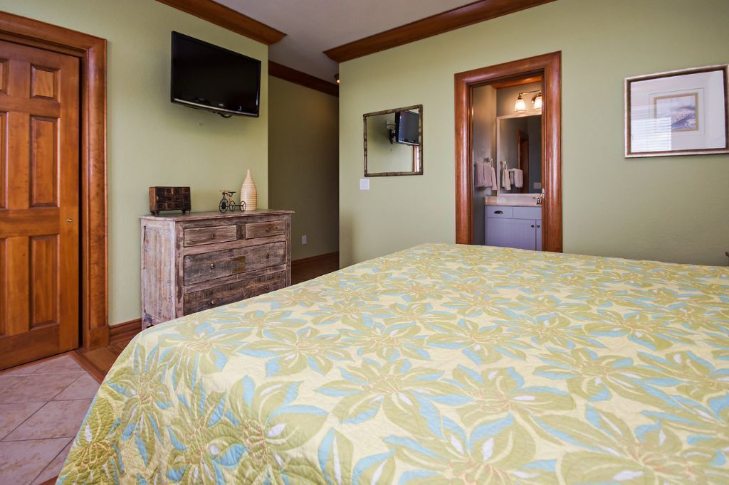 A bedroom with a floral bedspread, a wooden dresser, a television mounted on the wall, and a bathroom with a mirror and sink.