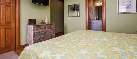 A bedroom with a floral bedspread, a wooden dresser, a television mounted on the wall, and a bathroom with a mirror and sink.