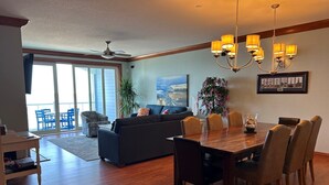 A spacious living room with a dark-colored sofa, a dining table with chairs, a chandelier, and a large window with a view. - WV25 Surf Shack by First Flight Rentals (Rodanthe)