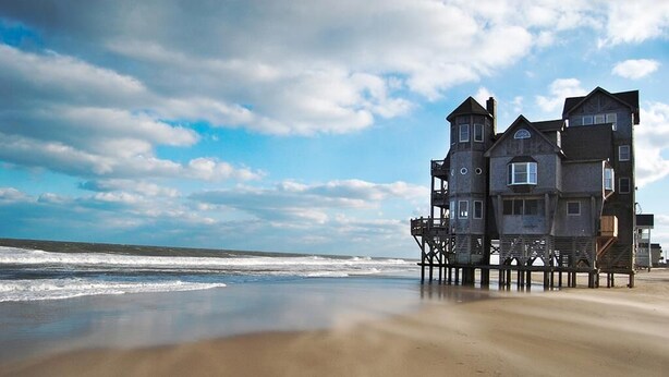 A coastal building with multiple levels and balconies, situated on a sandy beach with the ocean in the background. - WV25 Surf Shack by First Flight Rentals (Rodanthe)