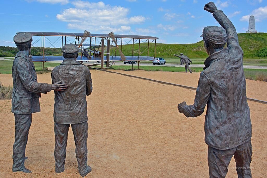 A statue of three figures in military uniforms standing in a line, with a biplane and a monument in the background.