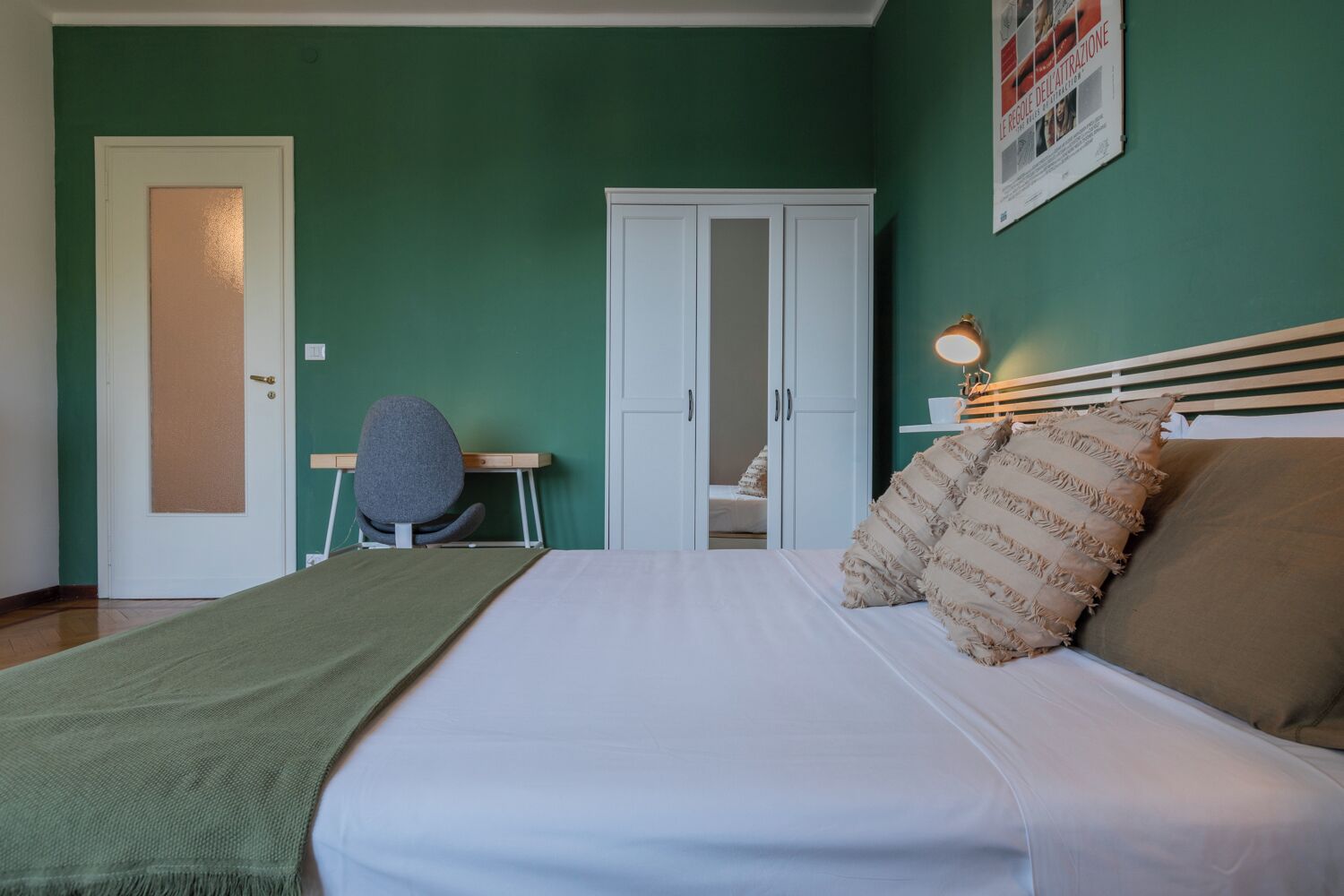A bedroom with a bed, a desk, a chair, a wardrobe, and a wall-mounted light.