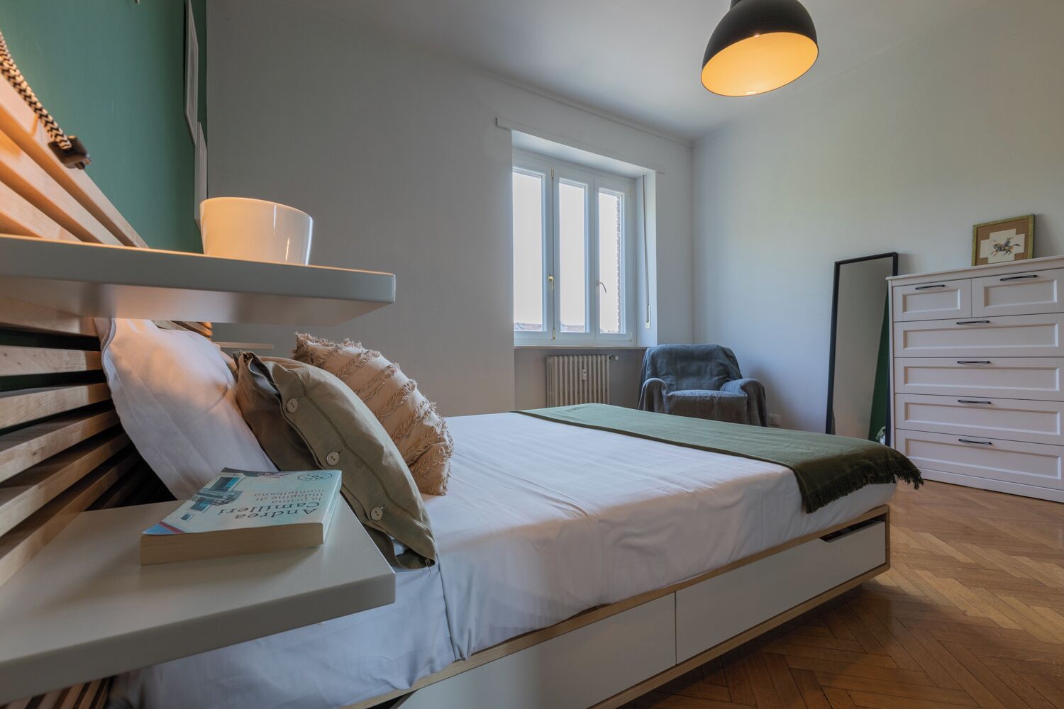 Un dormitorio con cama, mesita de noche, lámpara, silla, cómoda y ventana.