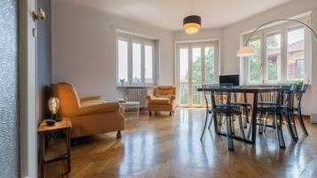Un salon spacieux doté d’un parquet en bois disposé en chevrons, d’un canapé, d’une table à manger avec des chaises et de grandes fenêtres laissant entrer la lumière naturelle.