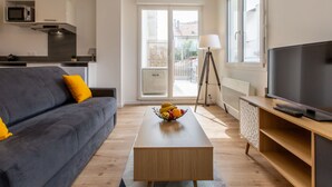 A modern living room with a grey sofa, a wooden coffee table with fruit, a TV on a wooden stand, and a kitchen in the background. - Cap Océan by Interhome (Capbreton)
