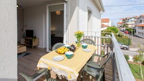 En balkong utomhus med ett matbord för två personer, en stol, ett litet bord med en vas med gula blommor och utsikt över gatan och de omkringliggande byggnaderna.