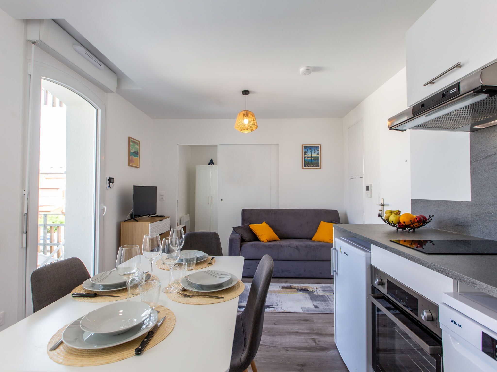 Eine kompakte Küche mit Einbauofen, ein Essbereich mit Tisch für vier Personen und ein graues Sofa mit orangefarbenen Kissen.