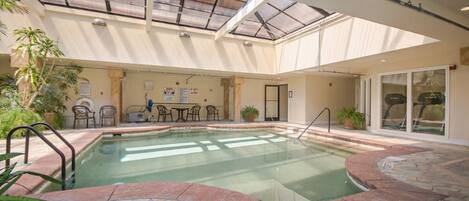 Una piscina interior con techo de vidrio, suelo de piedra y una zona de estar con mesas y sillas.