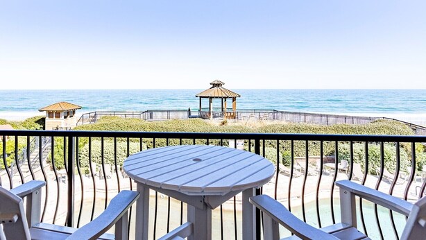 Un balcón con mesa y sillas, con vistas a una playa que incluye un pabellón y una torre de salvavidas.