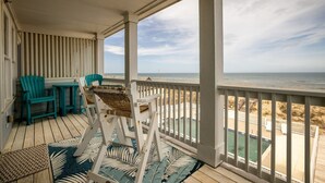 Área externa coberta com vista para a praia, apresentando um deck de madeira, duas cadeiras Adirondack turquesa, uma cadeira de balanço branca e um tapete tecido.