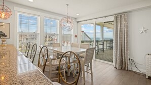 A modern dining area with a large table and chairs, a view of the ocean through large windows, and a balcony with a railing. - SC301- Langman's Sea Cabin by First Flight Rentals (Kill Devil Hills)