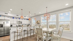A modern kitchen with white cabinetry, a central island with bar stools, pendant lighting, and large windows with blinds. - SC301- Langman's Sea Cabin by First Flight Rentals (Kill Devil Hills)