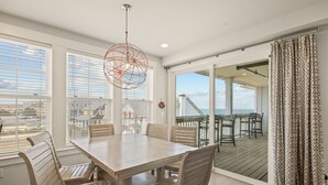 A dining area with a wooden table and chairs, large windows with blinds, a hanging light fixture, and a view of the city through the windows. - SC301- Langman's Sea Cabin by First Flight Rentals (Kill Devil Hills)
