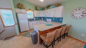 A compact kitchen with white cabinets, a central island with bar stools, a clock on the wall, and a window with blinds.