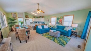 A living room with a blue sofa, a dining table with chairs, a ceiling fan, and a kitchen in the background.