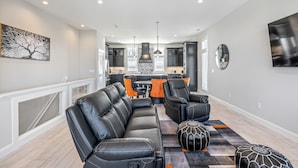 Een moderne woonkamer met een zwarte leren bank, een bijpassende fauteuil en een salontafel. Achtergrond is een keuken met donkere kasten en een groot raam.