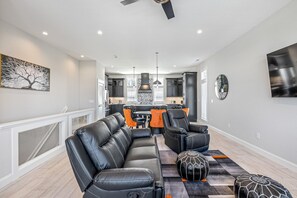 A modern living room with a black leather sofa, a matching armchair, and a coffee table. There is a kitchen in the background with dark cabinets and a large window.