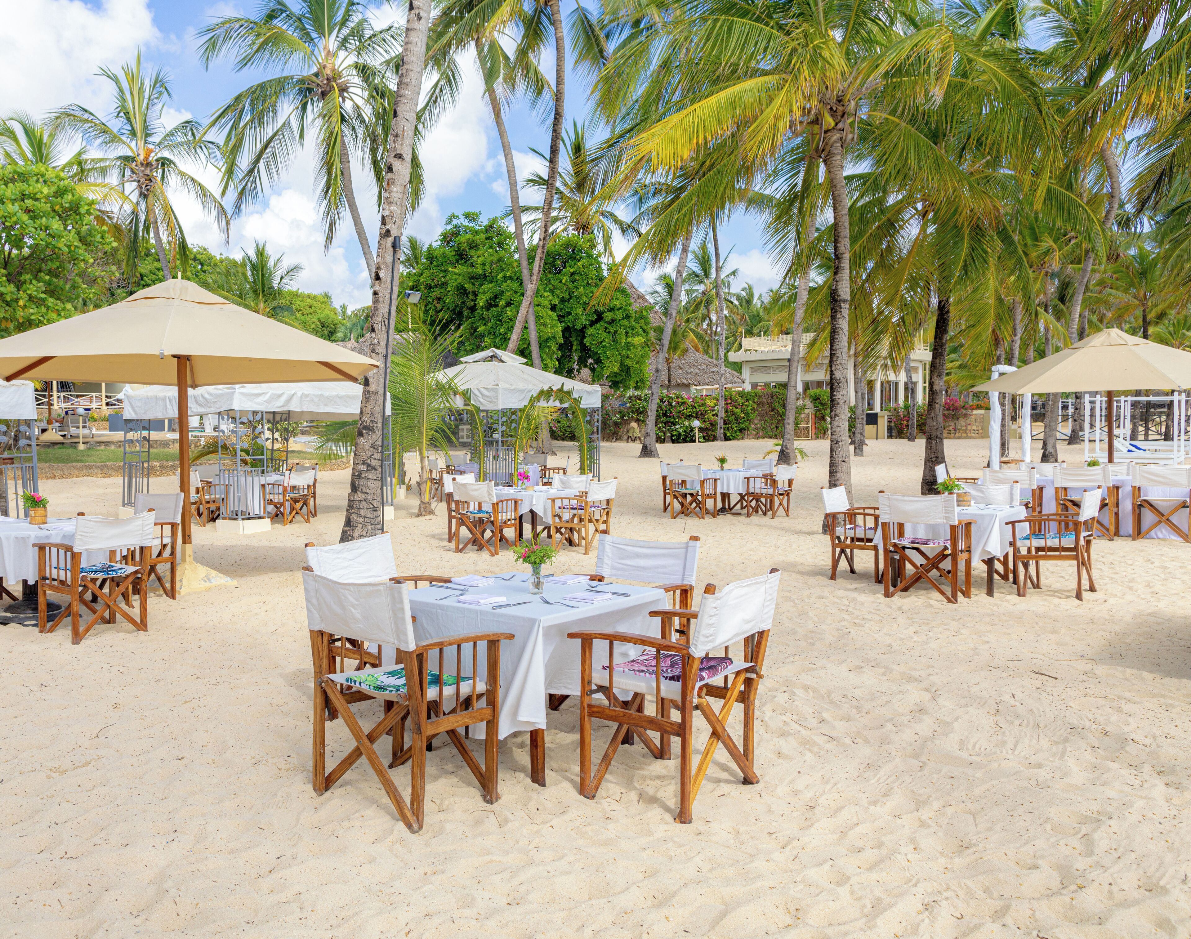 Un área de comedor al aire libre con mesas y sillas, rodeada de palmeras y sombrillas en una playa de arena.