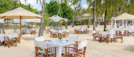 Un área de comedor al aire libre con mesas y sillas, rodeada de palmeras y sombrillas en una playa de arena.
