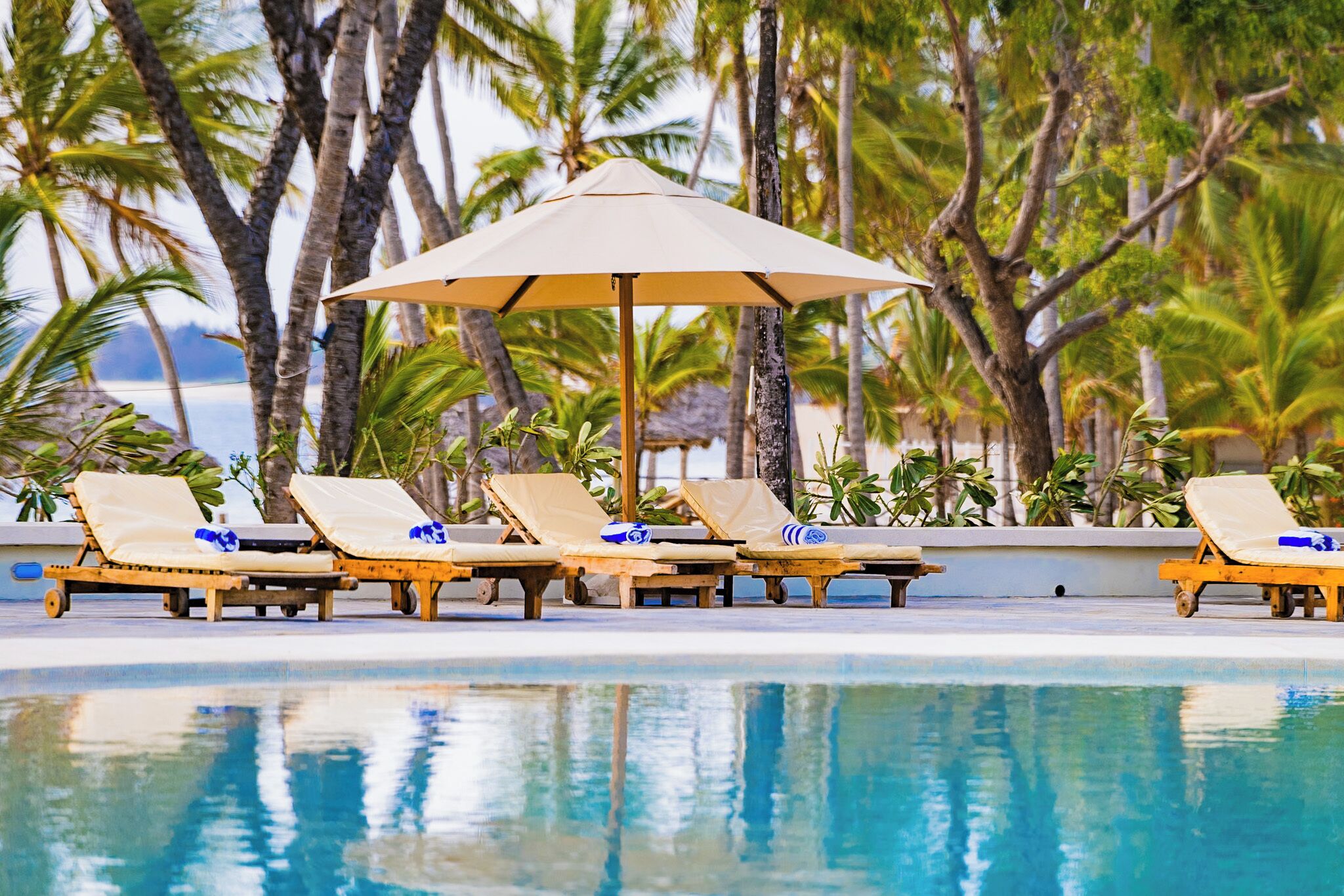 A poolside area with lounge chairs and an umbrella, surrounded by palm trees.
