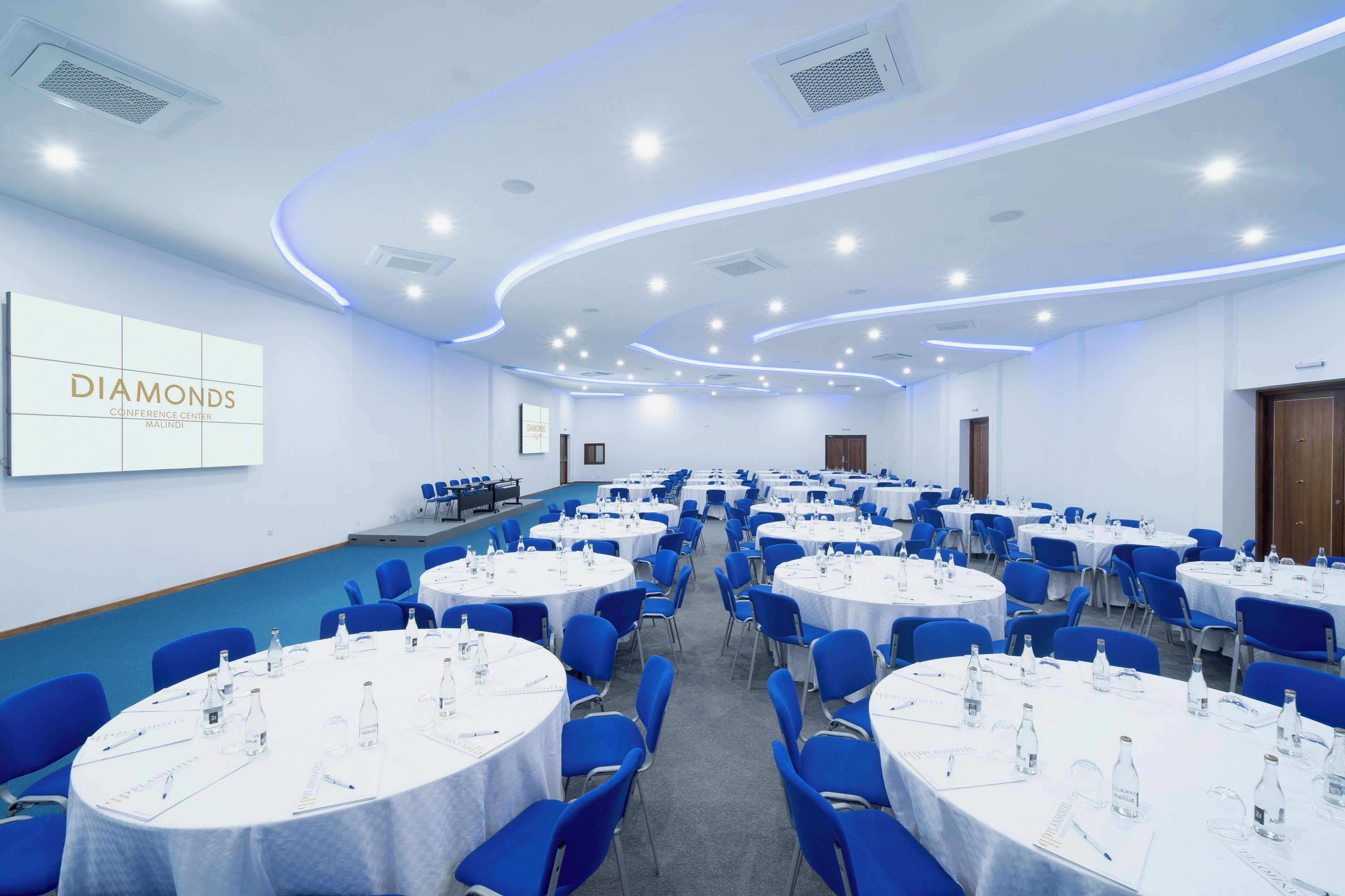 A conference room with round tables, blue chairs, and a large wall sign reading "DIAMONDS CONFERENCE CENTER MALINDI".