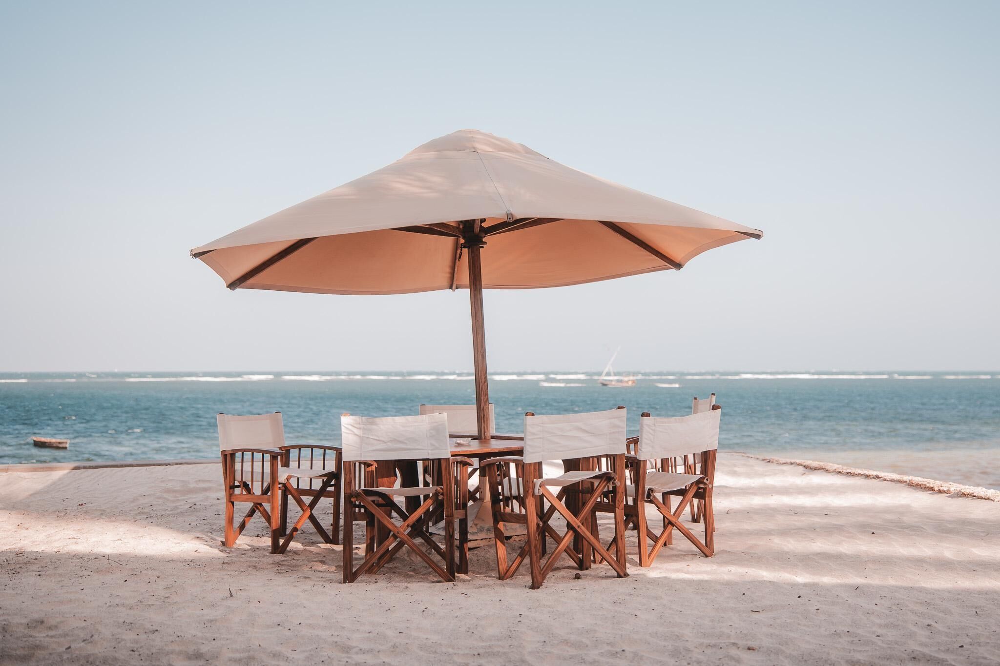 Une table installée sur la plage, protégée par un grand parasol, avec des chaises en bois et une vue sur l’océan.