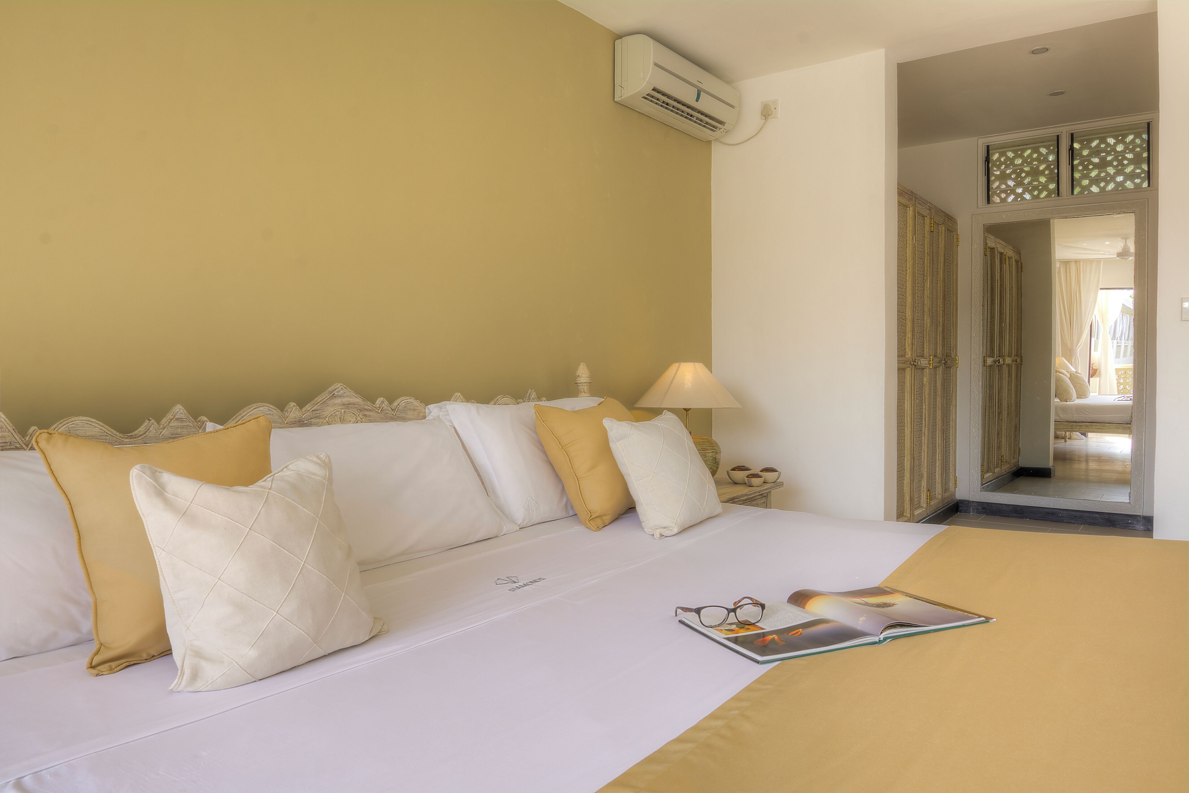 A neatly made bed with white and yellow pillows, a bedside lamp, and a magazine on the bed.