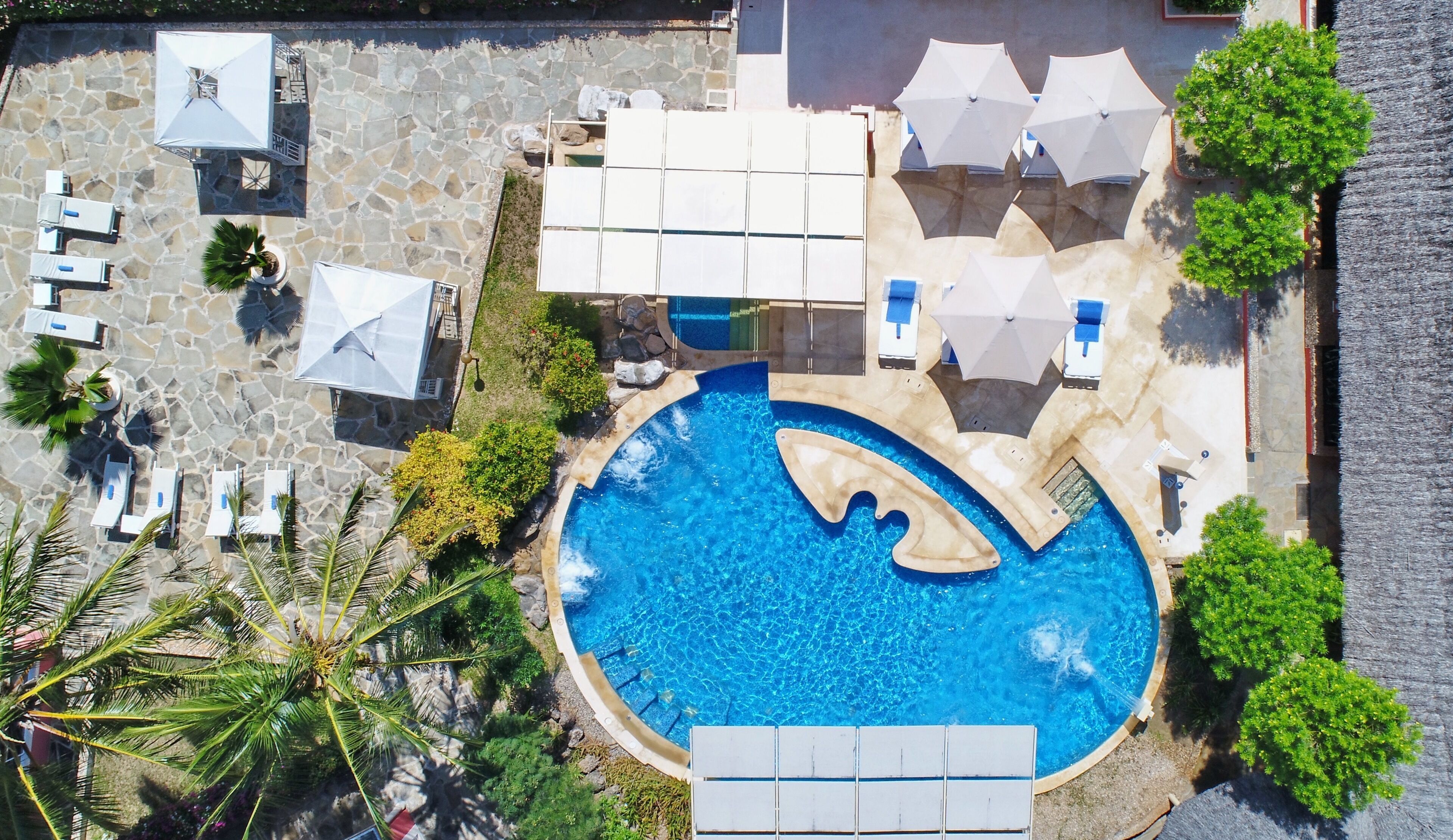 An aerial view of a resort with a swimming pool, lounge chairs, umbrellas, and palm trees.