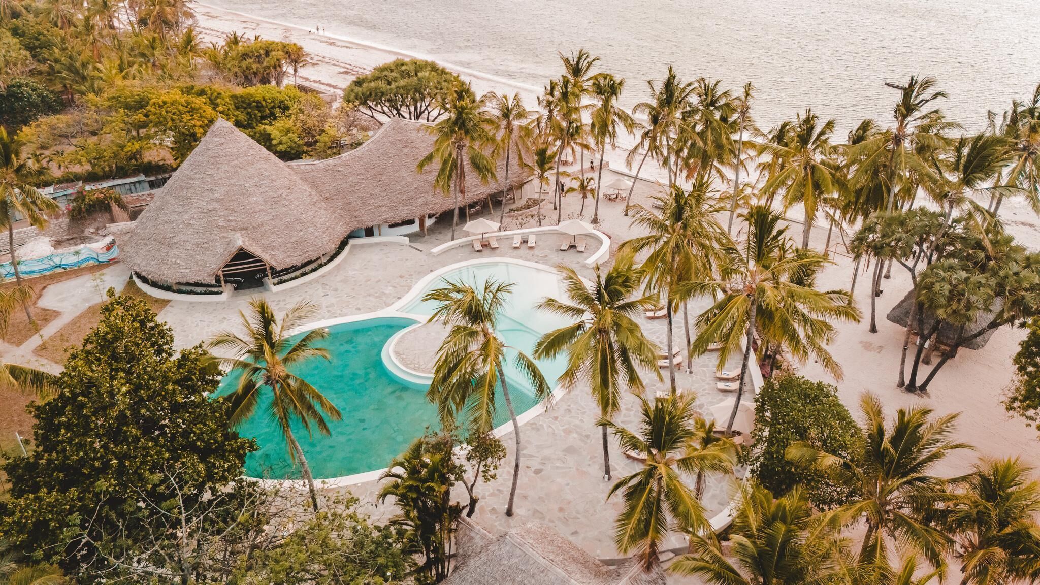 An aerial view of a resort with a central swimming pool, palm trees, and a thatched-roof building.