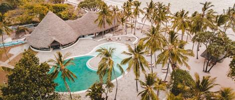 An aerial view of a resort with a central swimming pool, palm trees, and a thatched-roof building.