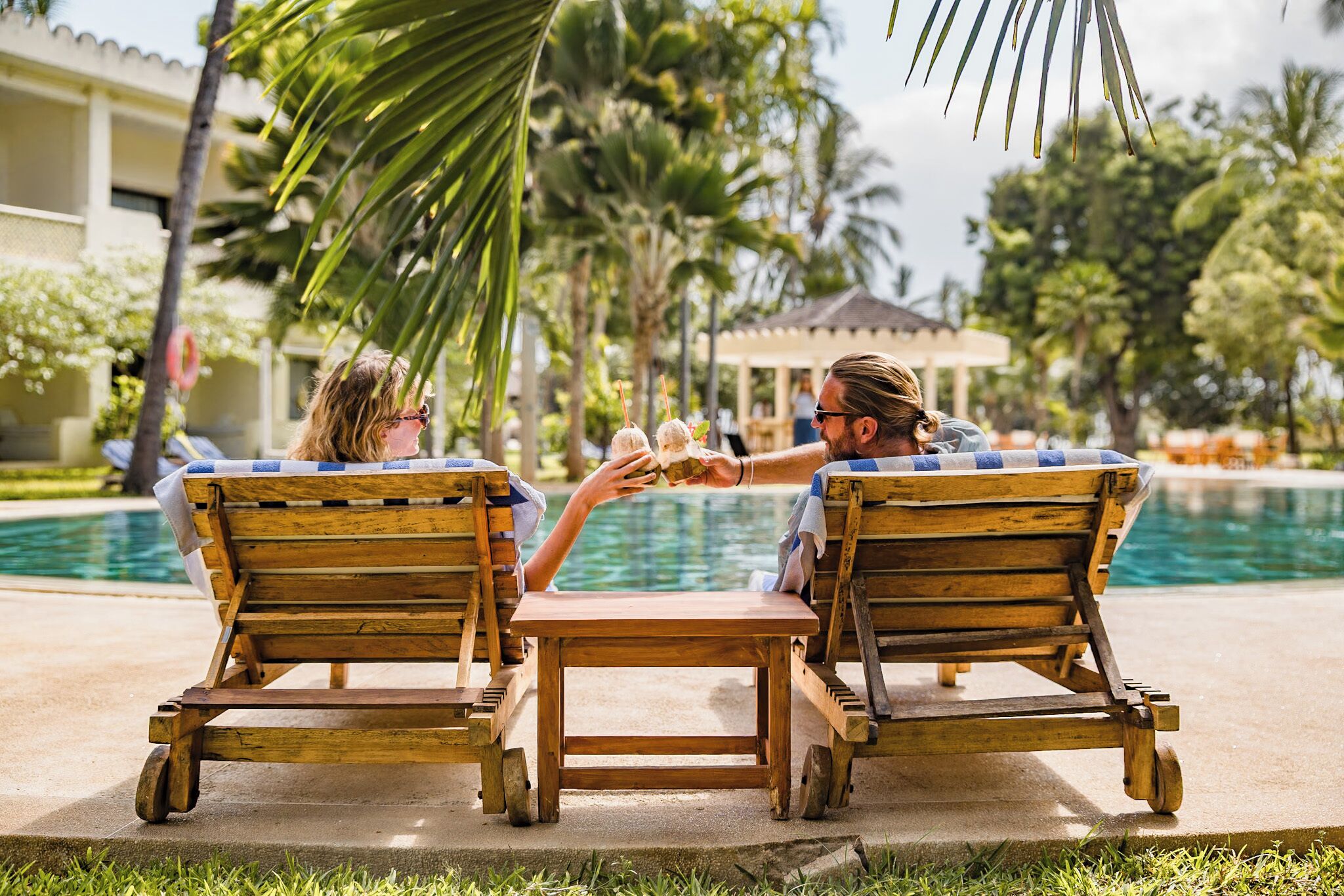 Two wooden lounge chairs with a small table between them, positioned by a poolside with palm trees and a building in the background.