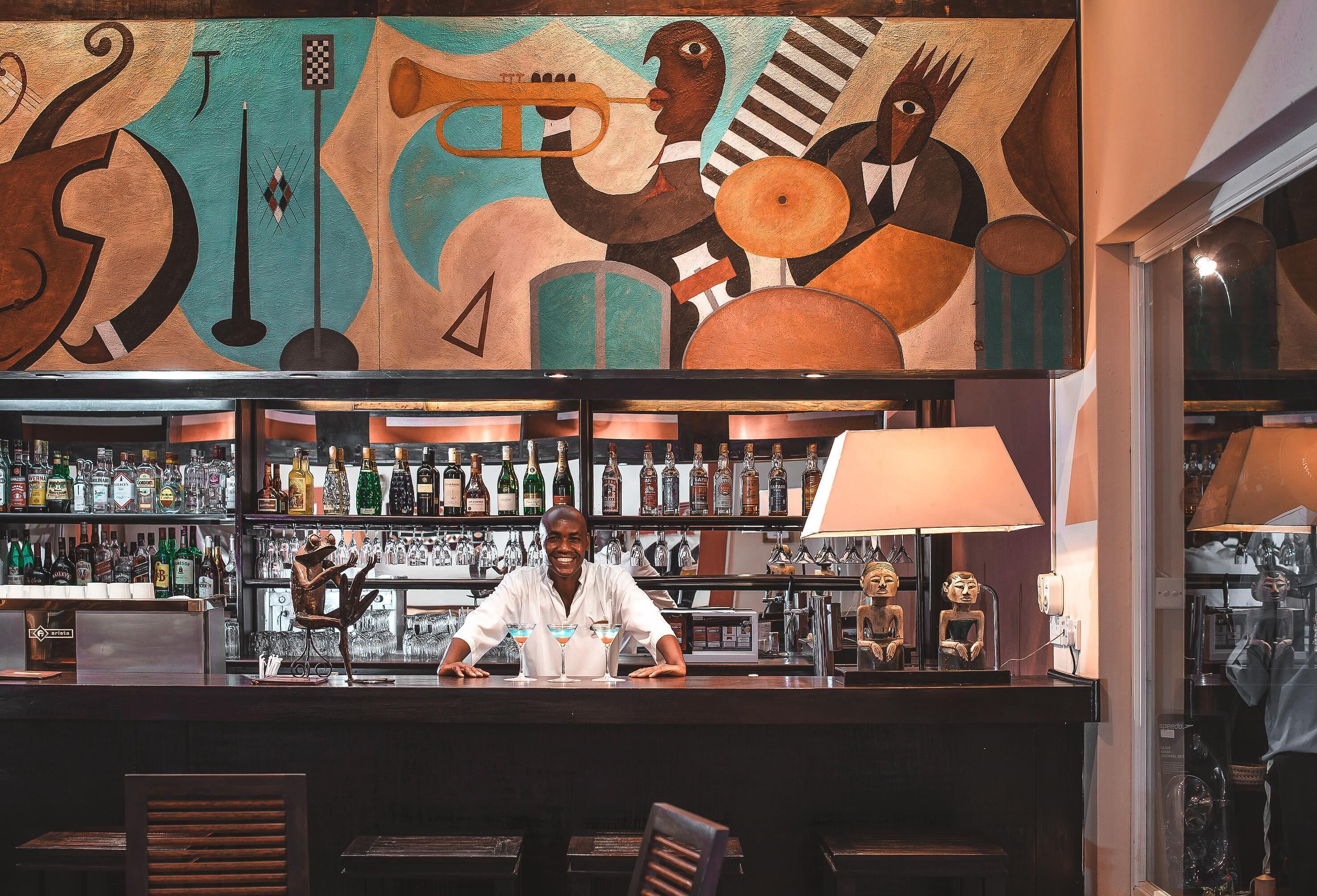 A bar area with a bartender, shelves of liquor bottles, and a mural on the wall.
