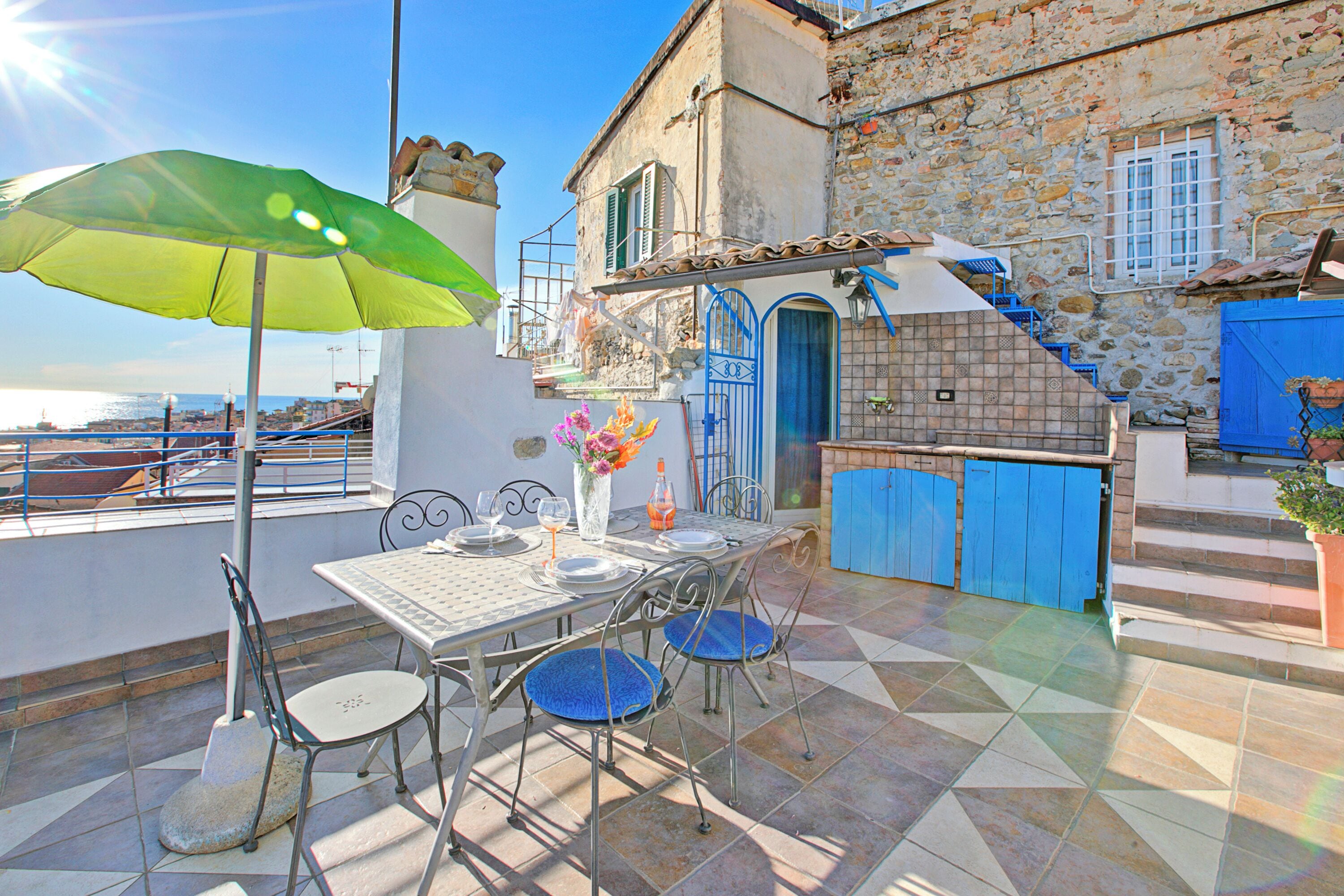 A terrace with a table set for dining, a green umbrella, and a stone building in the background.