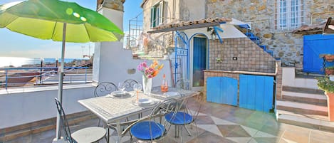 A terrace with a table set for dining, a green umbrella, and a stone building in the background.
