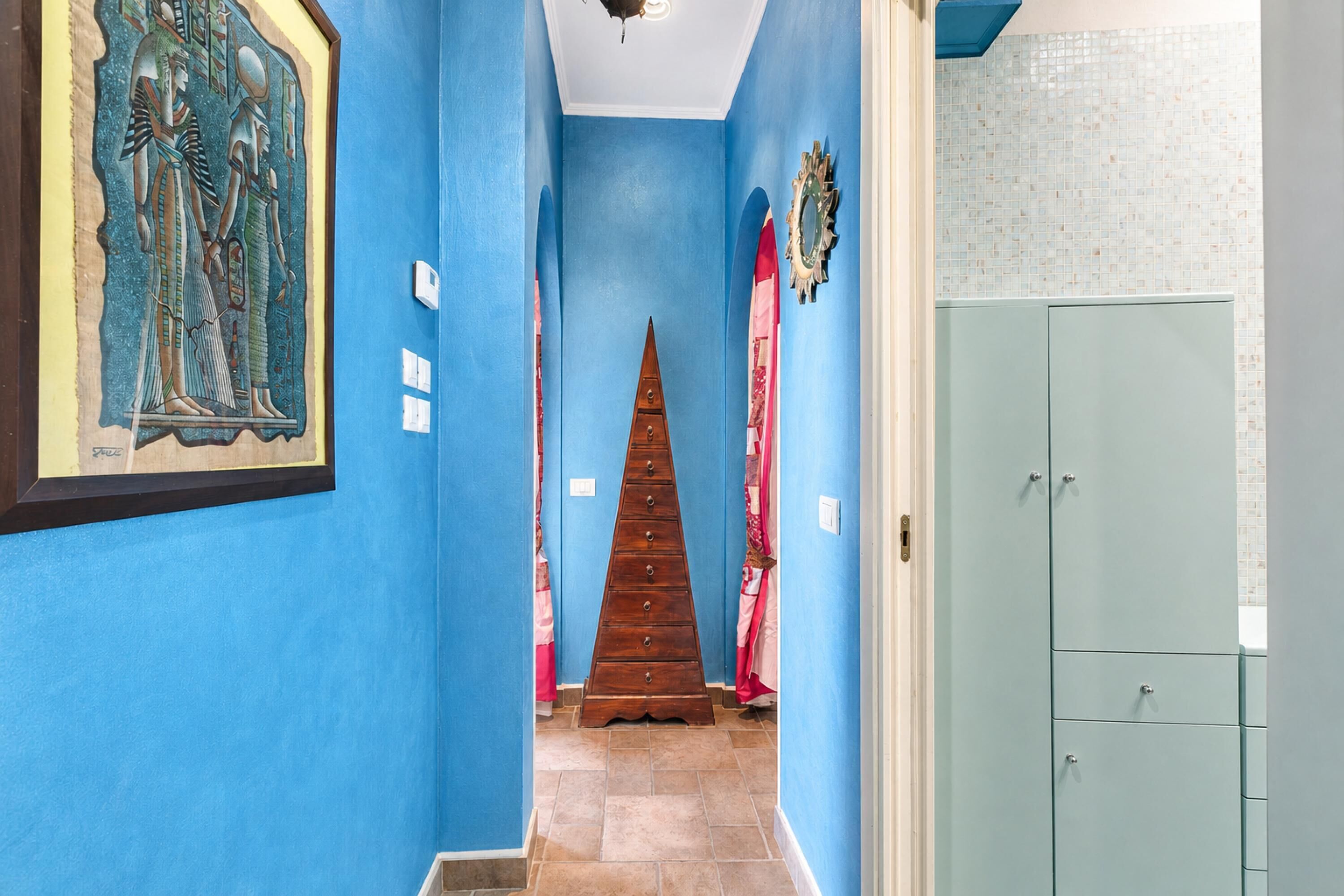 Un couloir aux murs bleus, une échelle en bois, un tableau ancien représentant une scène égyptienne encadré, et une salle de bain avec un mur carrelé.