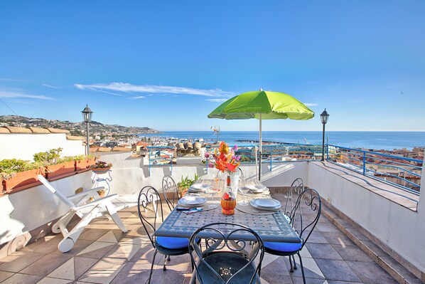 A terrace with a table set for dining, overlooking a coastal area with boats and buildings.