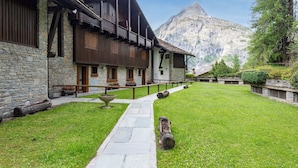 A stone building with wooden balconies, a stone pathway, and a mountain in the background. - Plan Gorret Wonderful Apartment In Courmayeur (Courmayeur)