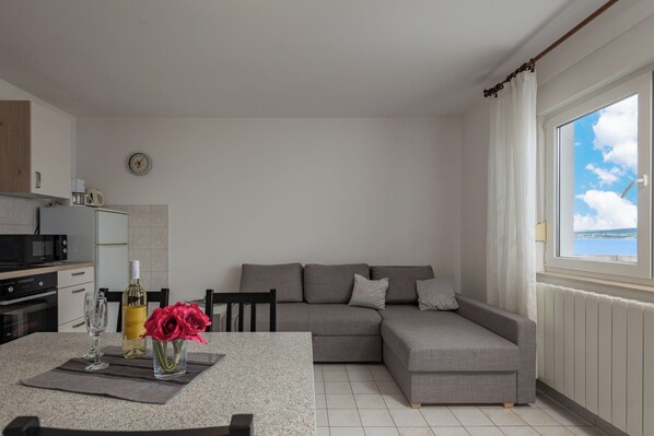 A modern kitchen with a dining area, a grey sofa, and a view of the sea through a window.