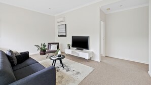 Uma sala de estar moderna com um sofá cinzento, uma mesa de centro, uma TV de tela plana em um suporte branco e um vaso com planta.